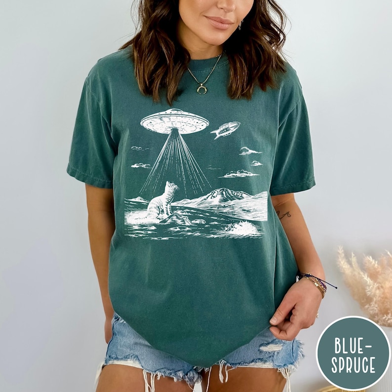 Pode incluir: Uma camiseta azul turquesa com um gr&aacute;fico branco de um gato olhando para cima para um OVNI. O OVNI est&aacute; emitindo raios de luz. O texto "Blue Spruce" &eacute; vis&iacute;vel no canto inferior direito da imagem.