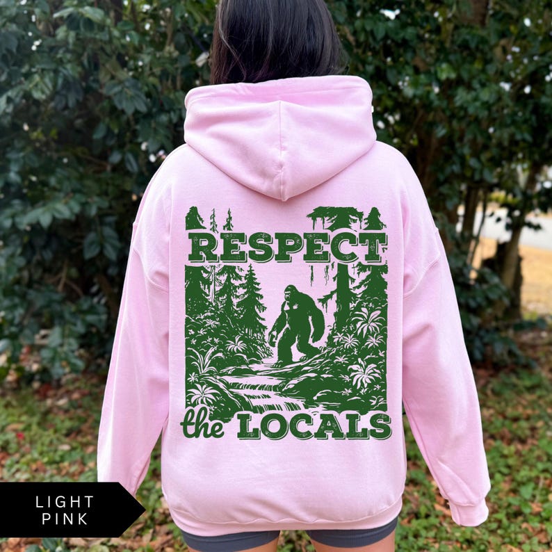 May include: Light pink hooded sweatshirt with a green graphic print of a Bigfoot in the woods. The graphic says "Respect the Locals".