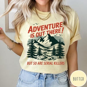 以下が含まれることがあります： 赤と黒で「ADVENTURE IS OUT THERE！BUT SO ARE SERIAL KILLERS」と書かれた黄色のTシャツ。グラフィックは山脈、木、鳥を描いています。Tシャツは明るい黄色です。