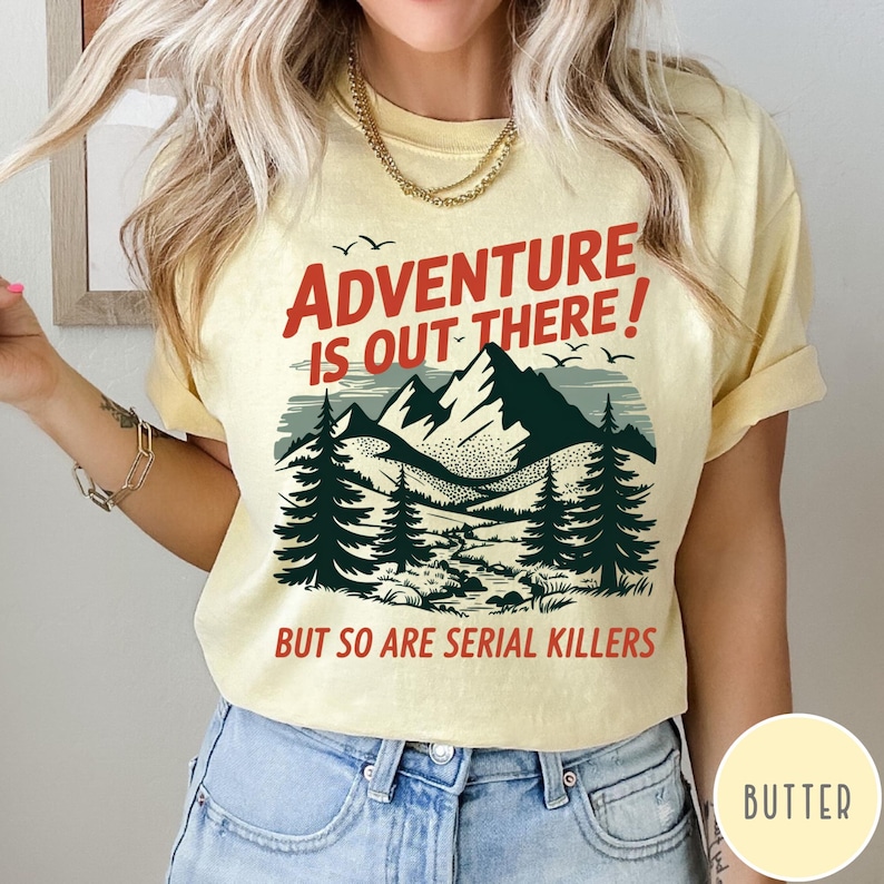 以下が含まれることがあります： 赤と黒で「ADVENTURE IS OUT THERE！BUT SO ARE SERIAL KILLERS」と書かれた黄色のTシャツ。グラフィックは山脈、木、鳥を描いています。Tシャツは明るい黄色です。