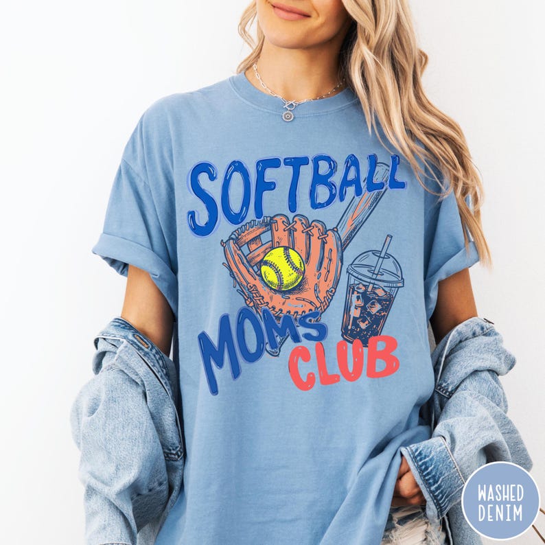 May include: Light blue t-shirt with the words "Softball Moms Club" in blue and red. The shirt features a softball glove with a yellow softball and a drink with ice. The shirt is made of washed denim.