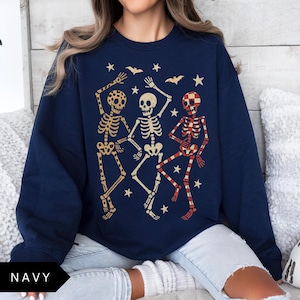 Puede incluir: Sudadera azul marino con tres esqueletos bailando en dorado y rojo, con estrellas y murci&eacute;lagos. La palabra "NAVY" est&aacute; en la parte inferior. Un dise&ntilde;o divertido y festivo para Halloween.