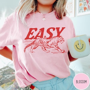 Może przedstawiać: Jasnor&oacute;żowa bluza z napisem "EASY" w czerwonych literach nad czerwoną grafiką tygrysa. Bluza ma okrągły dekolt. Biały kubek z ż&oacute;łtą uśmiechniętą buzią jest trzymany w dłoni.