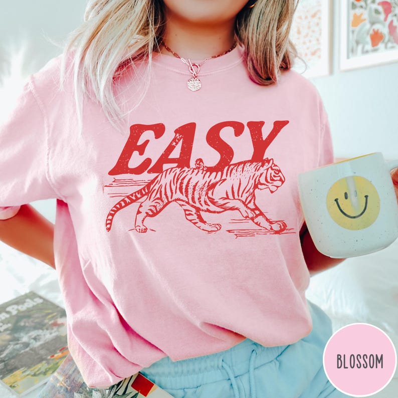 Może przedstawiać: Jasnor&oacute;żowa bluza z napisem "EASY" w czerwonych literach nad czerwoną grafiką tygrysa. Bluza ma okrągły dekolt. Biały kubek z ż&oacute;łtą uśmiechniętą buzią jest trzymany w dłoni.