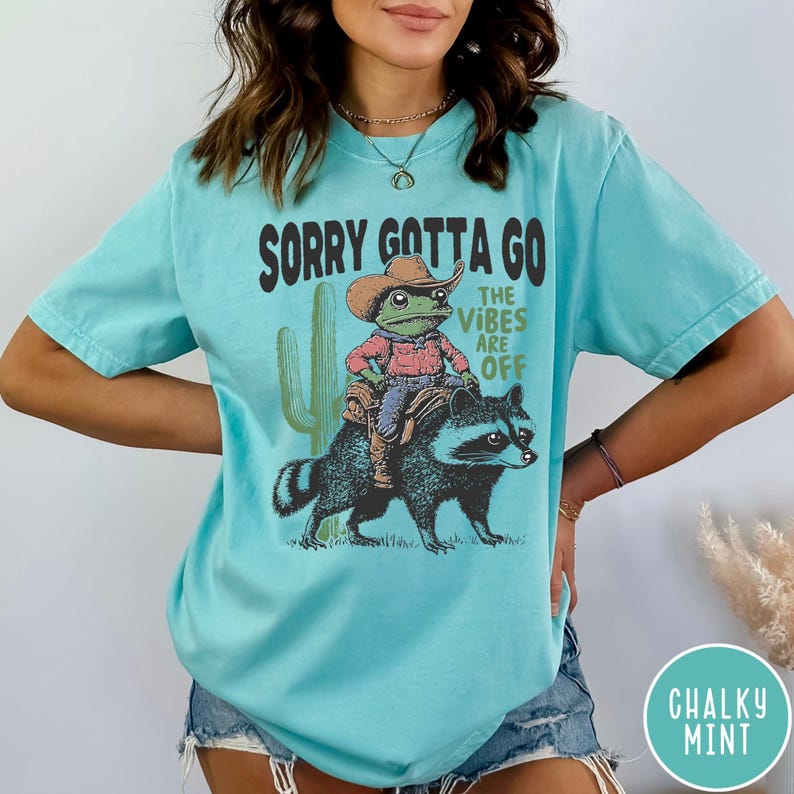 May include: A light blue t-shirt with the text "SORRY GOTTA GO THE VIBES ARE OFF." The graphic features a frog in a cowboy hat riding a raccoon, with a cactus in the background. The shirt has a relaxed fit and is made of a soft material.