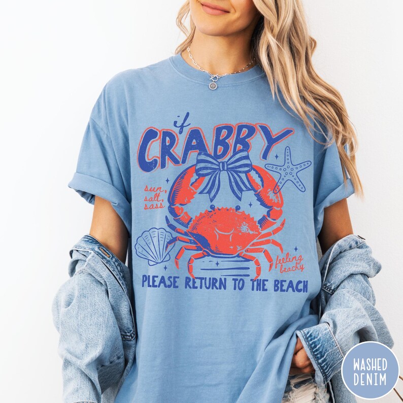 Pode incluir: Camiseta azul clara com um desenho de caranguejo vermelho com um la&ccedil;o azul, uma estrela do mar e uma concha. O texto diz "If Crabby, Please Return to the Beach". A camisa &eacute; de ganga lavada.