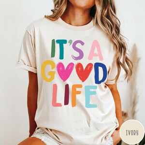K&ouml;nnte beinhalten: Ein elfenbeinfarbenes Kurzarm-T-Shirt mit dem bunten Schriftzug "IT'S A GOOD LIFE" auf der Vorderseite. Die Buchstaben und zwei Herzformen sind in verschiedenen leuchtenden Farben wie Gr&uuml;n, Lila, Pink, Gelb, Rot, Blau und Orange dargestellt.