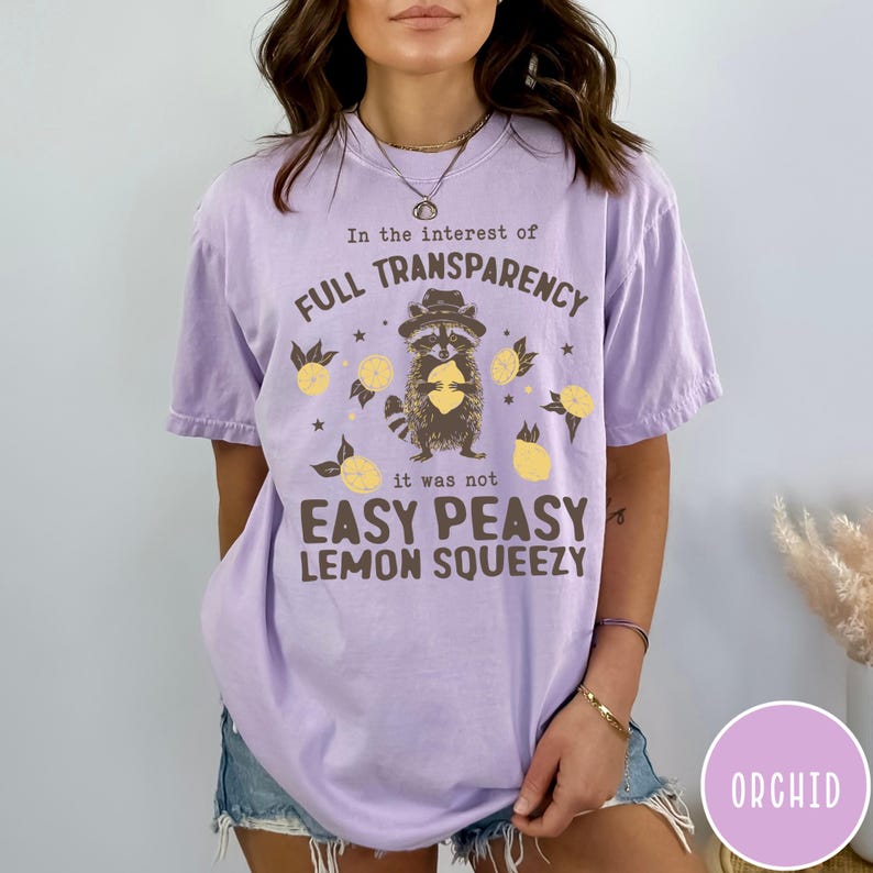 May include: A purple t-shirt with a raccoon wearing a hat and holding a lemon. The text on the shirt reads "In the interest of full transparency it was not easy peasy lemon squeezy".