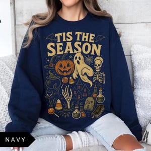 Puede incluir: Sudadera azul marino con un dise&ntilde;o de Halloween. El dise&ntilde;o incluye una calabaza, un fantasma, un esqueleto y el texto "TIS THE SEASON". Otros elementos incluyen un sombrero de bruja, caramelos y una casa embrujada.
