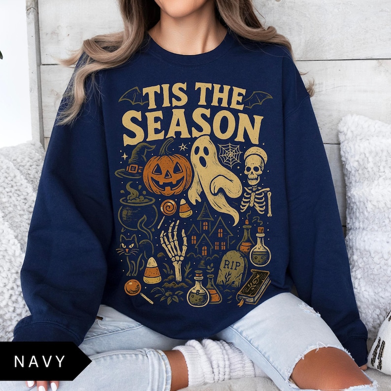 Puede incluir: Sudadera azul marino con un dise&ntilde;o de Halloween. El dise&ntilde;o incluye una calabaza, un fantasma, un esqueleto y el texto "TIS THE SEASON". Otros elementos incluyen un sombrero de bruja, caramelos y una casa embrujada.