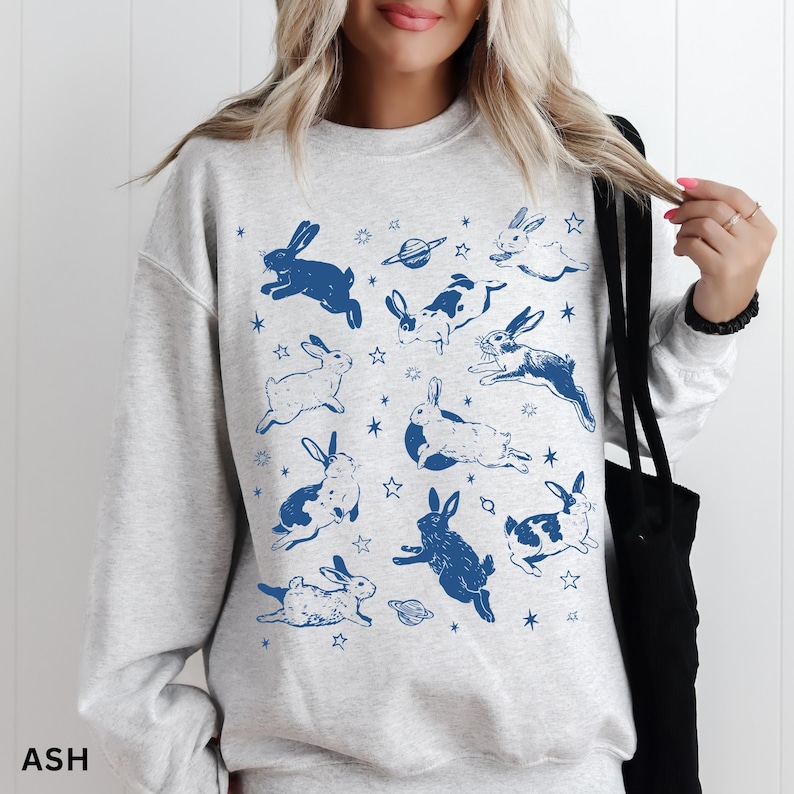 May include: Pale grey sweatshirt adorned with a pattern of blue rabbits and stars. The design includes illustrations of rabbits, planets, and stars. The word "ASH" is printed in the bottom left corner.