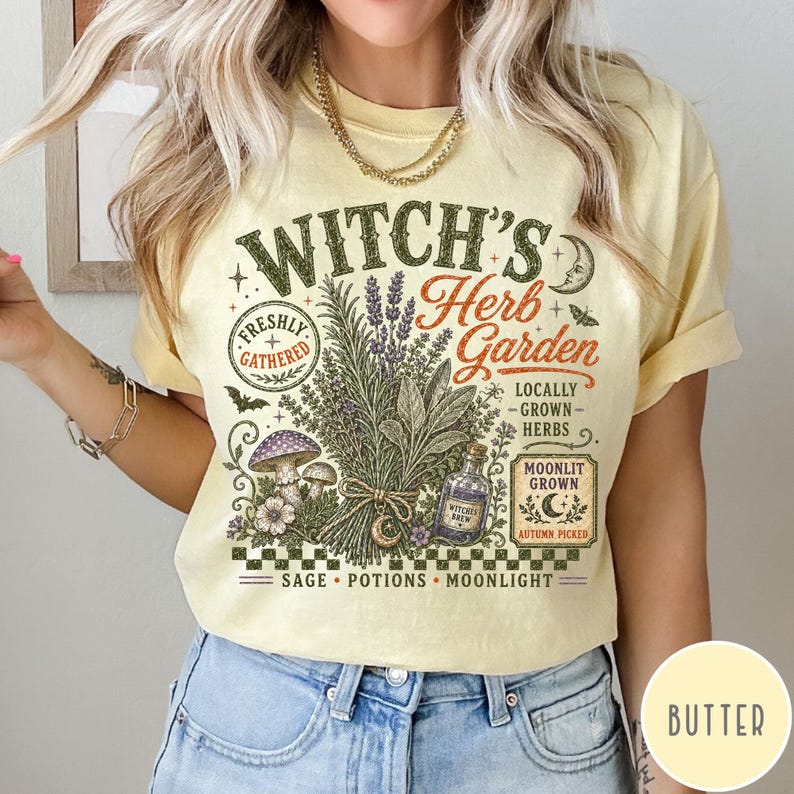 May include: A pale yellow t-shirt featuring a graphic print with the text Witch's Herb Garden. The design includes illustrations of a bundle of herbs, mushrooms, a potion bottle, a crescent moon, and the words Freshly Gathered and Moonlit Grown.