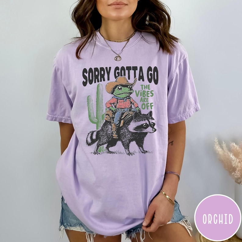 May include: A light purple t-shirt with a graphic of a frog in a cowboy hat riding a raccoon, with a cactus and the text "SORRY GOTTA GO THE VIBES ARE OFF". The shirt is a relaxed fit.