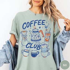 May include: A light green t-shirt with a "Coffee Club" graphic in blue and orange. The design includes a coffee pot, cups, and a glass of iced coffee. The words "all day, every day" are also included.