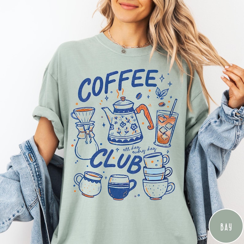 May include: A light green t-shirt with a "Coffee Club" graphic in blue and orange. The design includes a coffee pot, cups, and a glass of iced coffee. The words "all day, every day" are also included.
