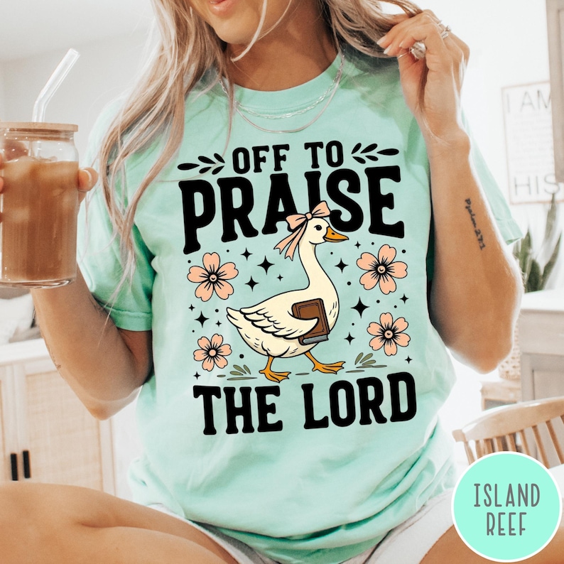 Puede incluir: Camiseta de color verde azulado claro con las palabras negras "OFF TO PRAISE THE LORD". En el centro, un ganso de dibujos animados con un lazo y un libro. Flores rosas y estrellas rodean al ganso.