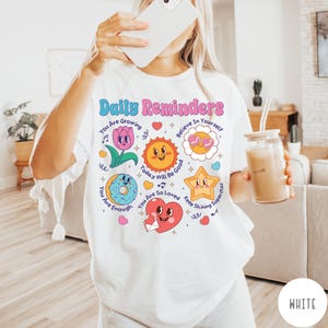May include: White t-shirt with the text "Daily Reminders" in pink and blue. The shirt features colorful illustrations of a flower, sun, donut, star, and heart with positive affirmations. The shirt is a comfortable, casual style.