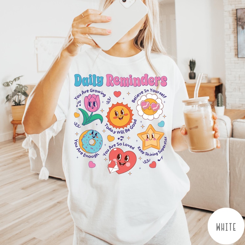 May include: White t-shirt with the text "Daily Reminders" in pink and blue. The shirt features colorful illustrations of a flower, sun, donut, star, and heart with positive affirmations. The shirt is a comfortable, casual style.
