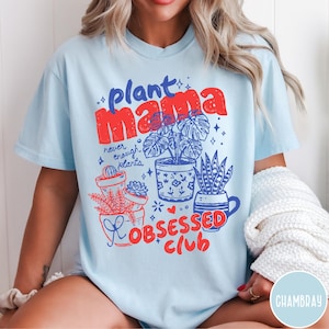 Puede incluir: Camiseta azul claro con las palabras "Plant Mama" en rojo y azul, junto con la frase "never enough plants". La camiseta presenta ilustraciones de plantas en macetas y las palabras "Obsessed Club" en rojo.