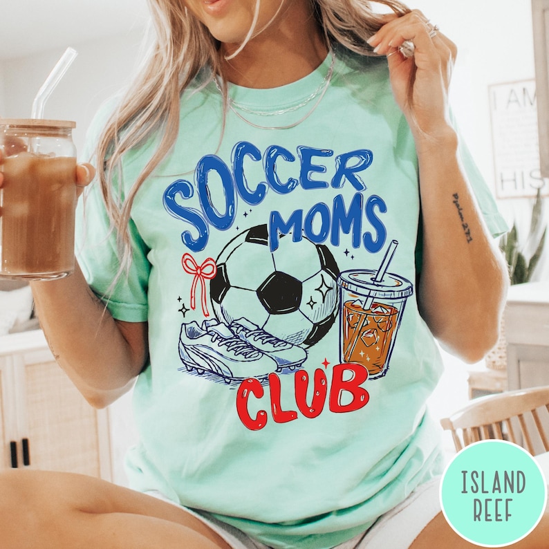 May include: A light green t-shirt with the words "Soccer Moms Club" in blue and red. The shirt features a soccer ball, cleats, and a drink with a straw. The person is holding a glass of iced coffee.