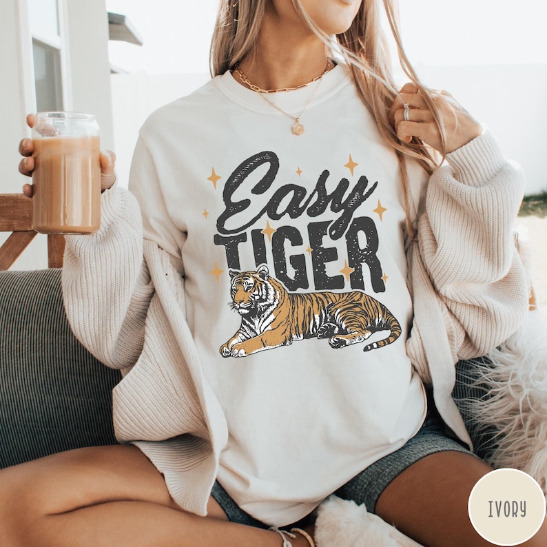 May include: A white sweatshirt with the text "Easy Tiger" in black lettering and a stylised tiger illustration. The tiger is orange and black with a white background. The sweatshirt is ivory in colour.
