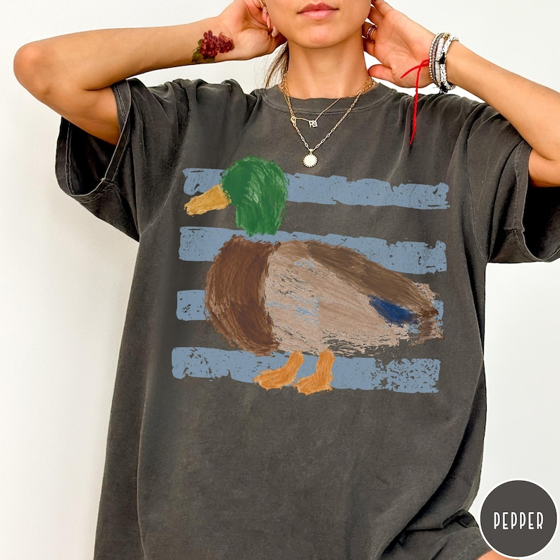 May include: A charcoal gray short-sleeve t-shirt featuring a painterly graphic of a mallard duck with a green head, brown body, and orange feet. The duck is positioned over three horizontal, distressed light blue stripes across the chest area.