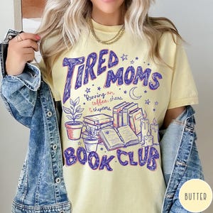 May include: Butter-colored t-shirt with a purple graphic that reads "Tired Moms Book Club." The design includes illustrations of books, candles, plants, and the text "Running on coffee, chaos & chapters."