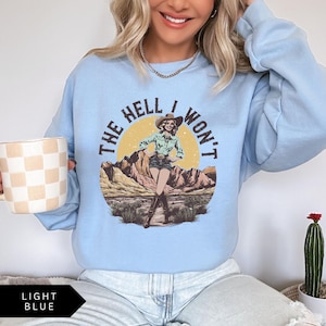 May include: Light blue sweatshirt with a graphic of a woman in a cowboy hat and the text "The Hell I Won't".