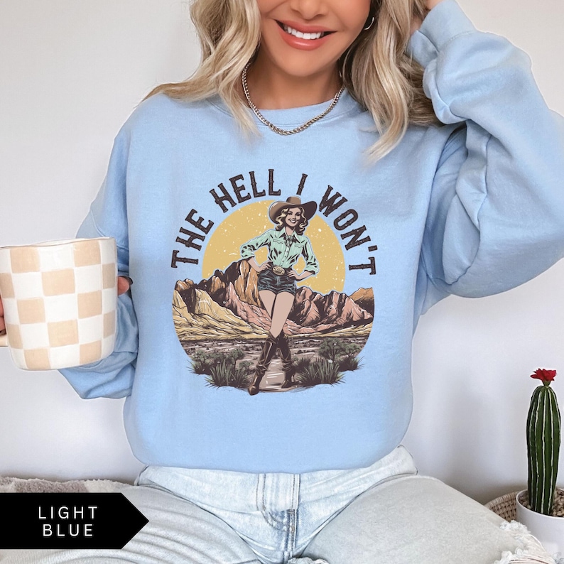 May include: Light blue sweatshirt with a graphic of a woman in a cowboy hat and the text "The Hell I Won't".