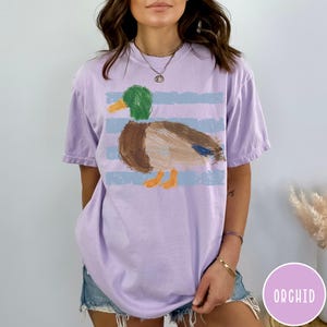 May include: A light purple short-sleeve t-shirt featuring a graphic of a mallard duck with a green head, brown body, and orange feet. The duck is positioned over horizontal light blue stripes. The shirt has a crew neckline and a relaxed fit.