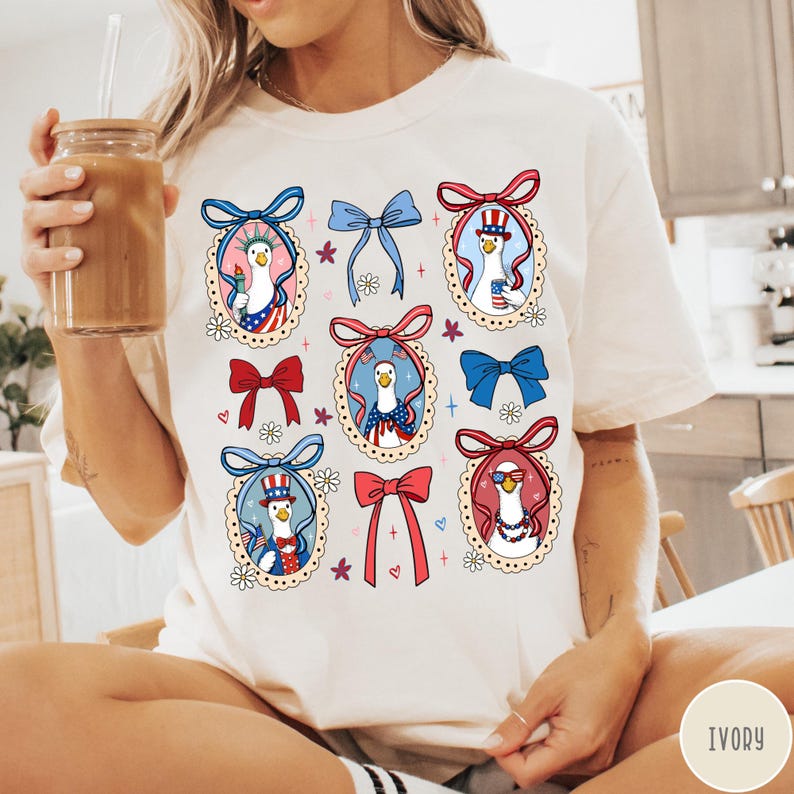 May include: Ivory-colored t-shirt featuring a repeating pattern of cartoon geese in patriotic attire, framed by decorative oval borders and bows in red, white, and blue. The geese are wearing hats, holding candles, and wearing sunglasses.