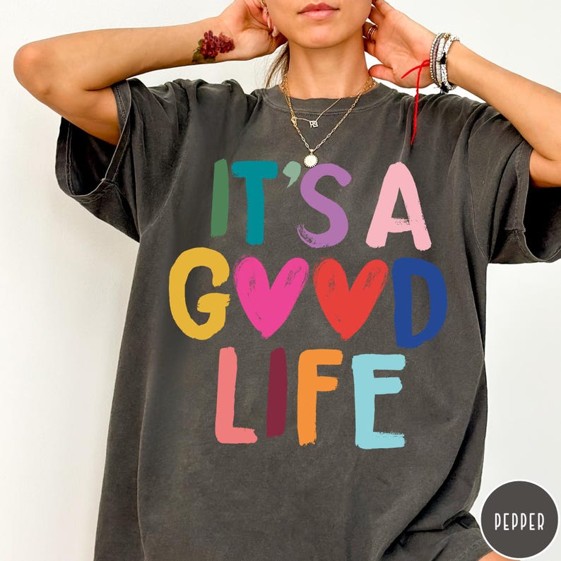 K&ouml;nnte beinhalten: Ein anthrazitfarbenes Kurzarm-T-Shirt mit dem bunten Schriftzug IT'S A GOOD LIFE, bei dem zwei Herzformen den Buchstaben O ersetzen. Die Buchstaben sind in verschiedenen Farben gedruckt, darunter Gr&uuml;n, Blau, Lila, Pink, Gelb, Rot und Orange.