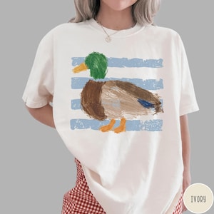 May include: An ivory short-sleeve t-shirt featuring a graphic of a mallard duck with a green head, brown body, and orange feet. The duck is layered over horizontal light blue distressed stripes. The shirt has a crew neckline and loose fit.