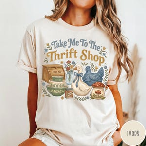 Op de afbeelding: Ivoor kleurig T-shirt met een grillige afbeelding. Het ontwerp bevat de tekst "Take Me To The Thrift Shop" boven illustraties van een brooddoos, kommen, een eend, een kip en een paddenstoel. Het shirt is een comfortabele, casual stijl.