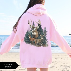 Op de afbeelding: Lichtroze hoodie met een gedetailleerde afbeelding van herten in een bos. Het ontwerp bevat een bok met grote geweien, twee kleinere herten en dennenbomen. De hoodie is effen lichtroze van kleur.