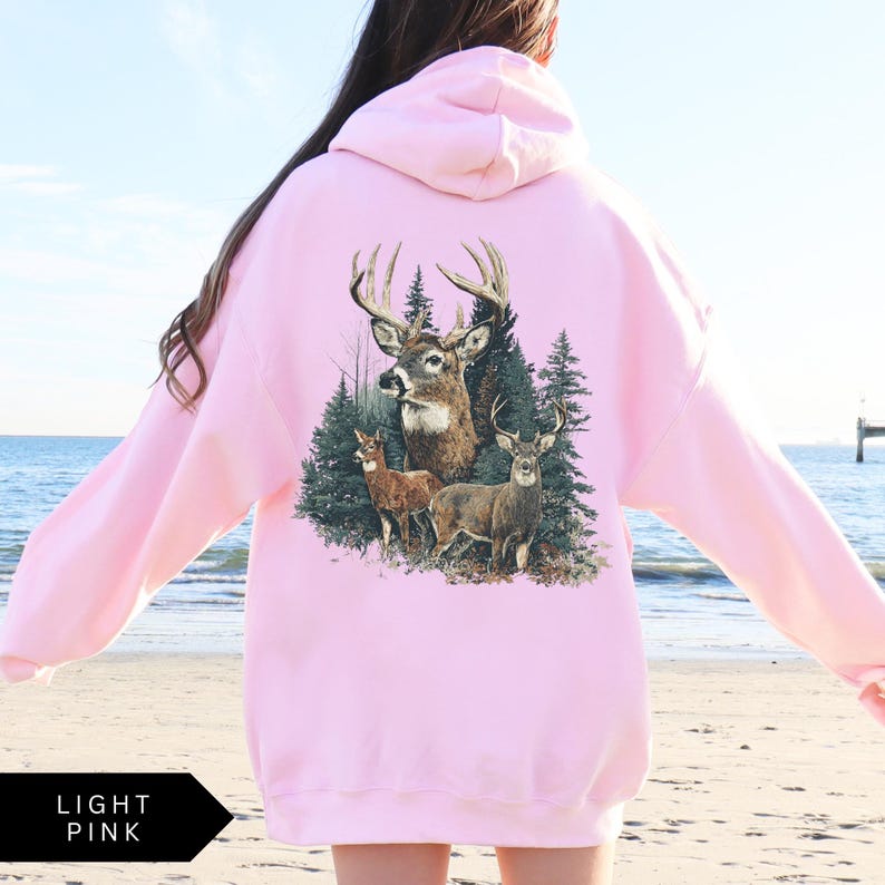 Op de afbeelding: Lichtroze hoodie met een gedetailleerde afbeelding van herten in een bos. Het ontwerp bevat een bok met grote geweien, twee kleinere herten en dennenbomen. De hoodie is effen lichtroze van kleur.
