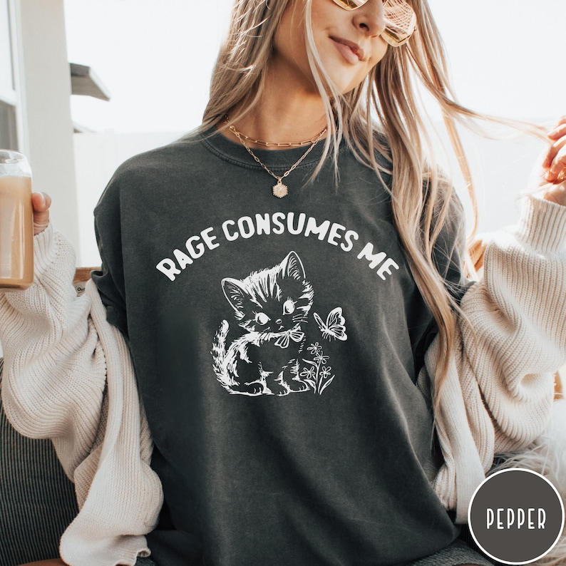 Puede incluir: Una sudadera gris oscuro con el texto "Rage Consumes Me" en letras blancas. Un gatito de dibujos animados blanco con una mariposa est&aacute; centrado en la sudadera.