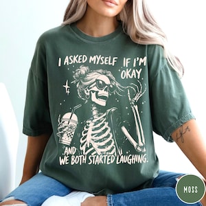 May include: Moss green t-shirt with a skeleton graphic wearing sunglasses and holding a drink. The text on the shirt reads, "I asked myself if I'm okay and we both started laughing."