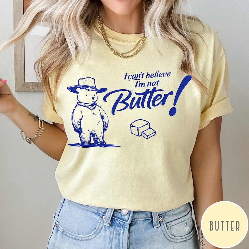 May include: A yellow t-shirt with a cartoon bear wearing a cowboy hat and the text "I can't believe I'm not Butter!" in blue. A stick of butter is also pictured.