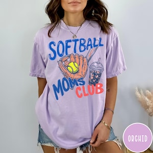May include: A light purple t-shirt with the words "SOFTBALL MOMS CLUB" in blue and coral. The graphic includes a softball glove, a yellow softball, and a drink with ice. The shirt is a comfortable, casual style.