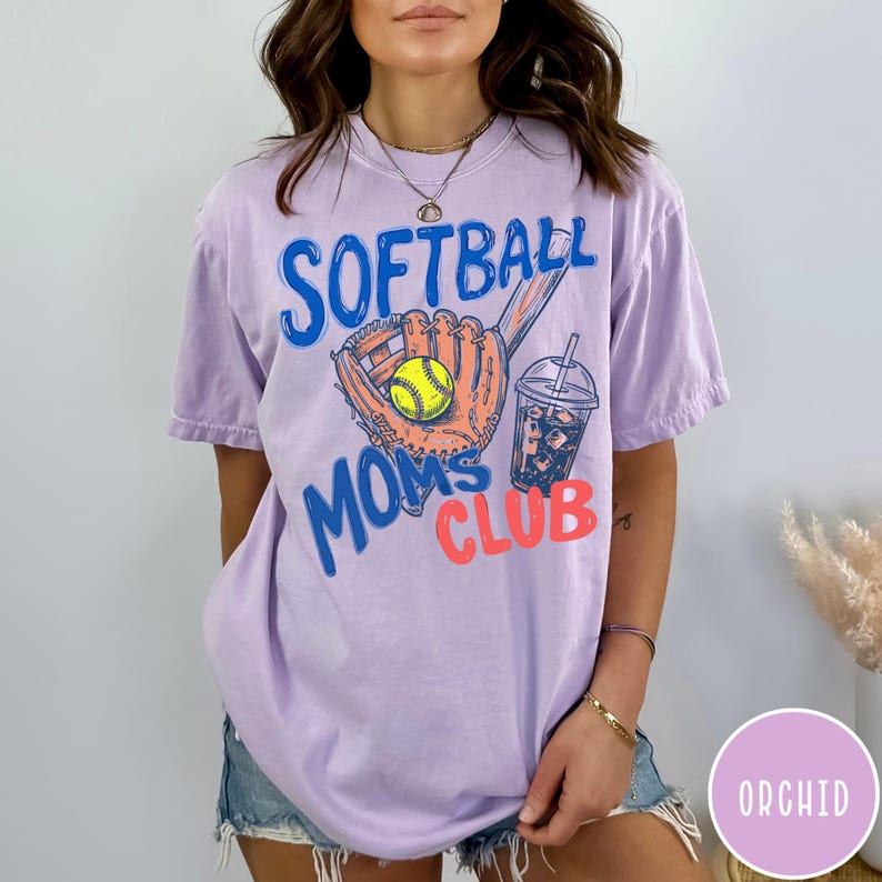 May include: A light purple t-shirt with the words "SOFTBALL MOMS CLUB" in blue and coral. The graphic includes a softball glove, a yellow softball, and a drink with ice. The shirt is a comfortable, casual style.