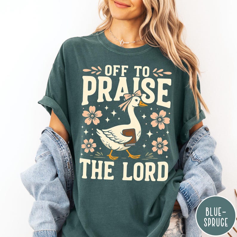 Puede incluir: Camiseta verde oscuro con el texto "OFF TO PRAISE THE LORD" en letras color crema. Un dibujo animado de un ganso con un libro y un lazo est&aacute; centrado debajo del texto, rodeado de flores rosas y estrellas. La camiseta es de corte holgado.
