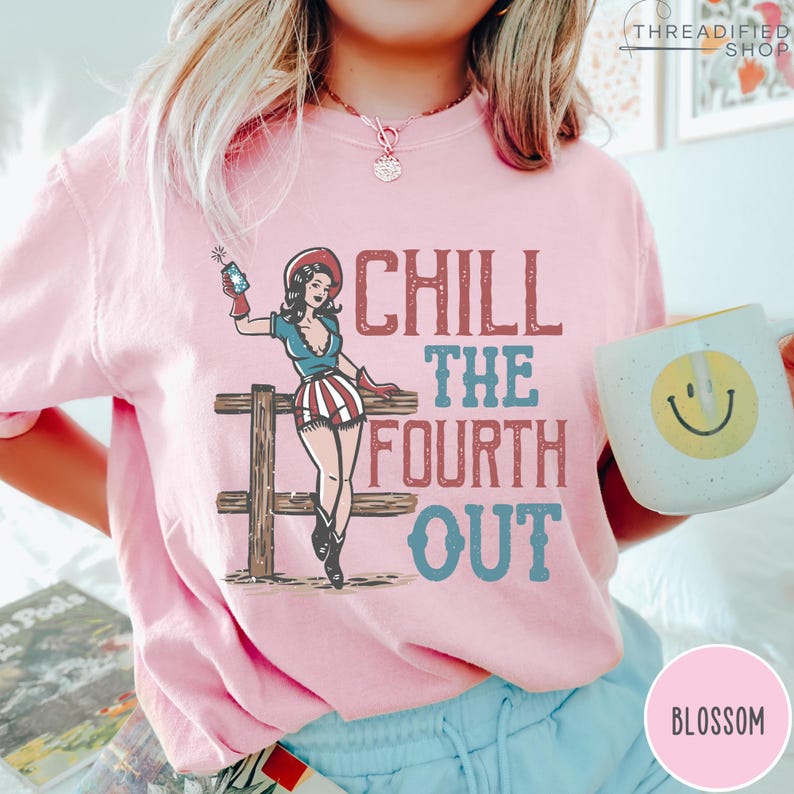 May include: A light pink sweatshirt with the text "CHILL THE FOURTH OUT" in red and blue. The graphic features a pin-up girl with a firework, a wooden fence, and a coffee mug with a smiley face.