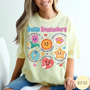 May include: A light yellow t-shirt with the words "Daily Reminders" in pink and blue. The shirt features cartoon graphics of a flower, sun, donut, star, and heart with positive affirmations. The shirt is made of a soft material.