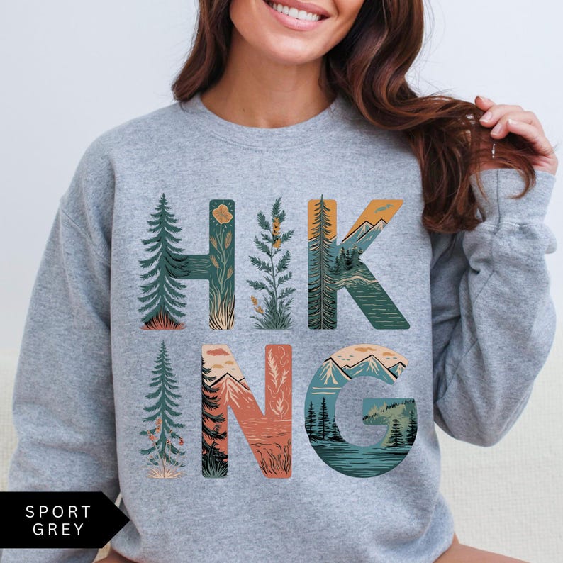 Puede incluir: Sudadera gris deportiva con la palabra "HIKING" en un dise&ntilde;o colorido con tem&aacute;tica de la naturaleza. Las letras est&aacute;n llenas de ilustraciones de &aacute;rboles, monta&ntilde;as y un lago. La sudadera tiene cuello redondo y mangas largas.