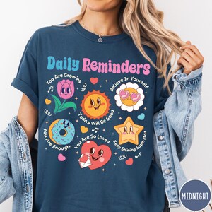 May include: Navy blue t-shirt with the text "Daily Reminders" and various cartoon graphics, including a flower, sun, donut, star, and heart. The shirt has positive affirmations like "You Are Enough" and "Believe in Yourself."