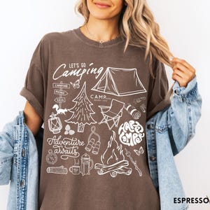 Może przedstawiać: Brązowa koszulka z białym motywem kempingowym. Projekt zawiera namiot, ognisko, drzewa i napisy "Let's Go Camping", "Adventure Awaits" i "Happy Camper".