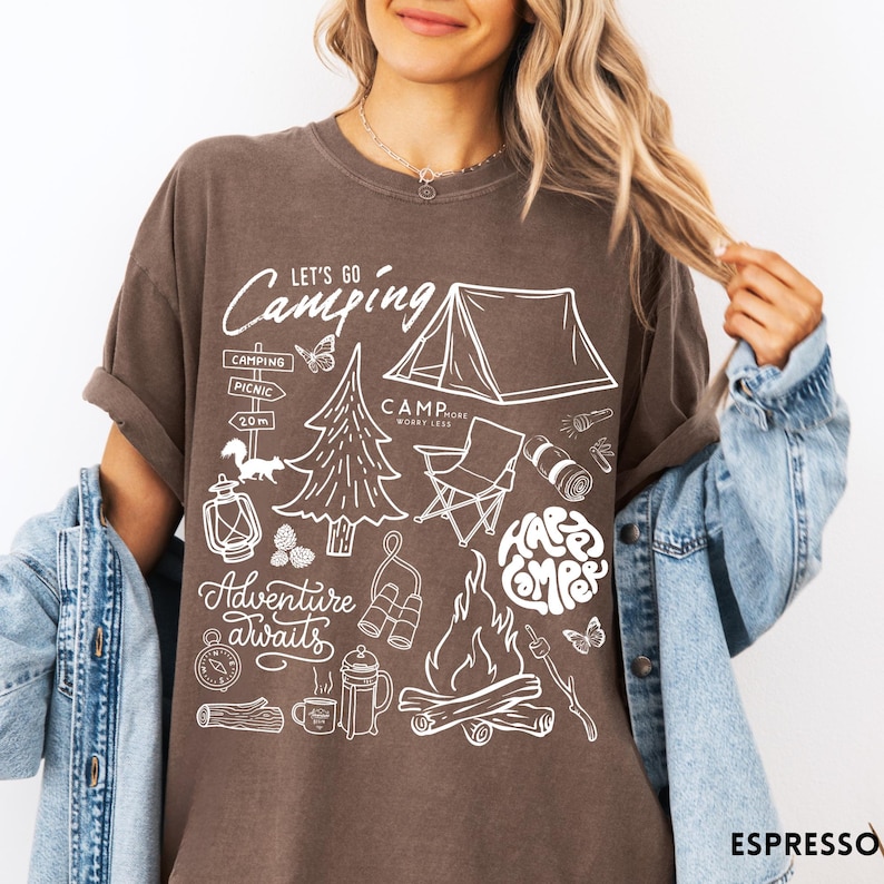 Może przedstawiać: Brązowa koszulka z białym motywem kempingowym. Projekt zawiera namiot, ognisko, drzewa i napisy "Let's Go Camping", "Adventure Awaits" i "Happy Camper".