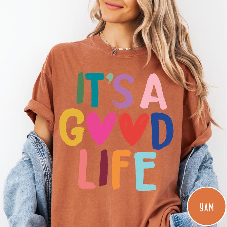 K&ouml;nnte beinhalten: Ein rostfarbenes Kurzarm-T-Shirt mit dem Text IT'S A GOOD LIFE in mehrfarbigen Blockbuchstaben. Die Buchstaben sind gr&uuml;n, blaugr&uuml;n, lila, rosa, gelb, rot, dunkelblau und hellblau, wobei zwei Herzformen den Buchstaben O ersetzen.