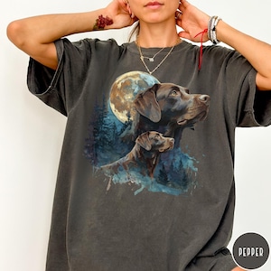 May include: Dark grey t-shirt featuring a watercolor illustration of two Labrador Retrievers with a full moon and forest backdrop. The design includes shades of brown, blue, and white.